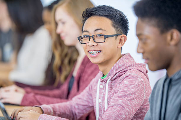 High School Students in Class A multi-ethnic group of high school age students are sitting in a row and are working in the computer lab on digital tablets. One man is smiling and looking at the camera. teenagers only teenager multi ethnic group student stock pictures, royalty-free photos & images