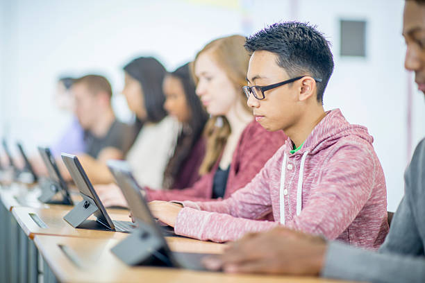 Students Sitting at Their Desks A multi-ethnic group of high school age students are sitting in a row and are working in the computer lab on digital tablets. secondary school activities stock pictures, royalty-free photos & images