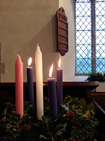 Advent candles in the traditional combination of three purple, one pink and one white candle in a wreath of holly, with a wooden hymn board hanging behind.