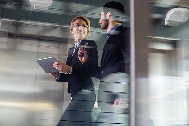 Two Business coworkers walking along elevated walkway Business professionals having a conversation while passing through office building lobby. on the move stock pictures, royalty-free photos & images