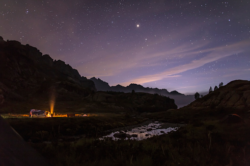 Tourists and hunters are heated around the campfire at night in the mountains in the background of the starry sky and fog