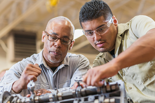 A mature African American man and young Hispanic man in an auto repair shop working together on a gasoline engine.  They are wearing a gray and beige shirts looking down at the engine with serious expressions.
