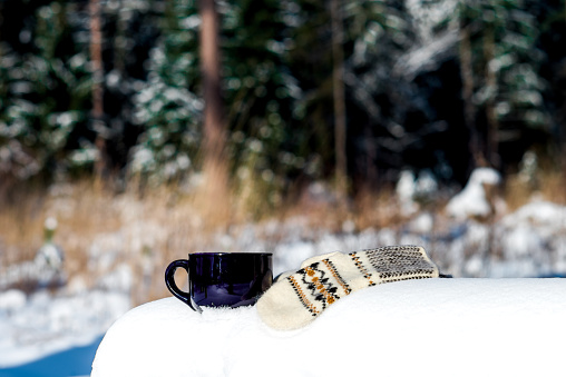 Cup and mittens on a snowy bench on the edge of winter forest.
