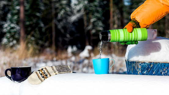 Female hand pouring water from thermos to the cup on a snowy bench on the edge of winter forest.