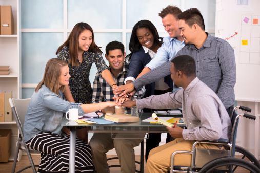 All hands in. Teamwork, unity. Creative young adults collaborate on business project together. Small business start up.  Smart casual office. One worker in wheelchair. 