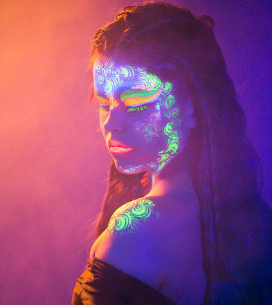 Portrait in half turn of beautiful young woman witn dreadlocks hairstyle. The girl is posing with fluorescent makeup on face and body. Female image prepared using a glowing neon cosmetics. Studio shooting with light haze in the UV light