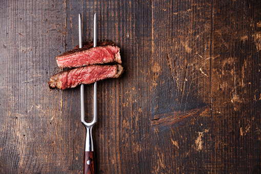 Slices of Medium rare grilled Steak Ribeye on meat fork on dark wooden background