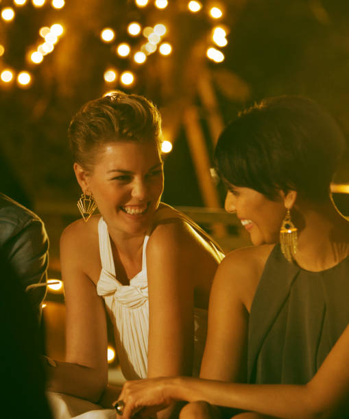 Three diverse young adult friends smiling and embracing during a fun night out, standing close together with an urban cityscape illuminated in the background