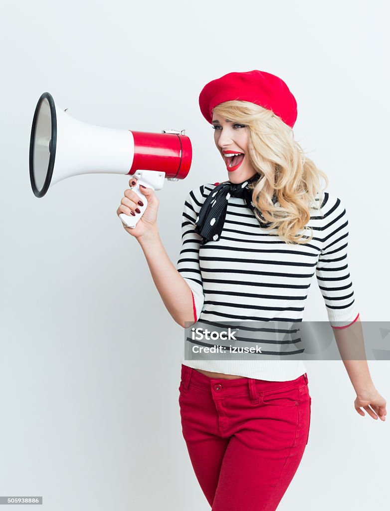 Blonde french woman wearing red beret shouting into megaphone Portrait of excited beautiful blonde woman in french outfit, wearing a red beret, striped blouse and neckerchief, shouting into megaphone. Megaphone Stock Photo