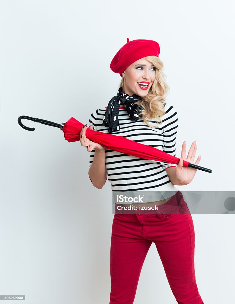Excited blonde french woman wearing red beret, holding an umbrella Portrait of excited beautiful blonde woman in french outfit, wearing a red beret, striped blouse and neckerchief, holding a red umbrella. Adult Stock Photo