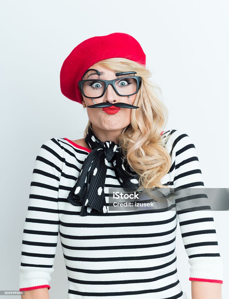 Playful blonde french woman wearing red beret and facial mask Portrait of playful blonde woman in french outfit, wearing a red beret, striped blouse, neckerchief and funny facial mask, staring at camera. Beret Stock Photo