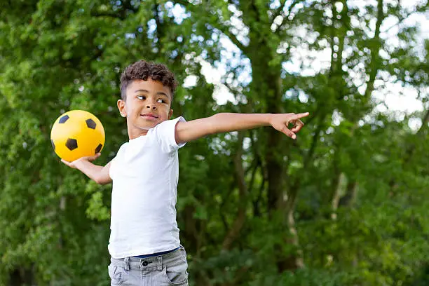 A happy little boy smiles as he gets ready to throw his yellow football. He is standing in his garden with lush green trees in the background.