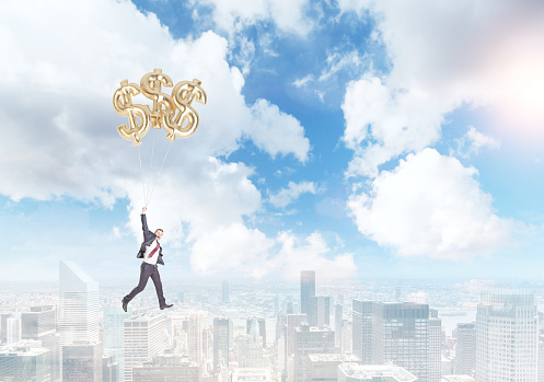 Businessman in a suit flying happily holding three balloons in shape of dollars over Paris, bright blue sky at the background. Concept of success and career growth