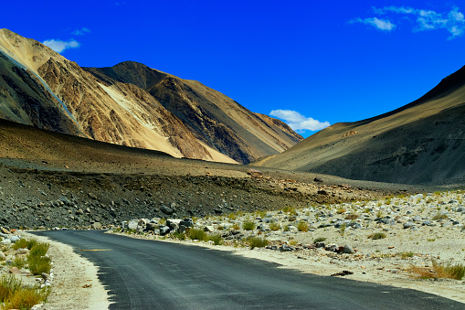 A concrete road towards beautiful rocky mountains and blue sky with peaks of Himalaya, Leh, Ladakh, Jammu and Kashmir, India