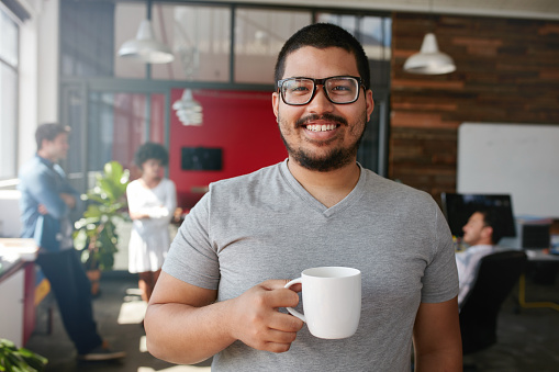 Portrait of smiling office worker having a coffee with his colleagues talking in the background.