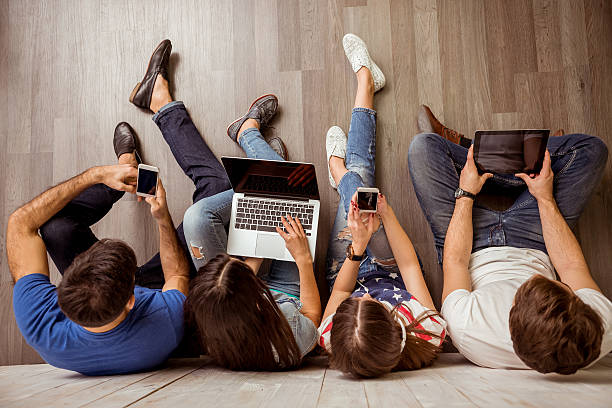 Group of young people Group of attractive young people sitting on the floor using a laptop, Tablet PC, smart phones, headphones listening to music, smiling digital native stock pictures, royalty-free photos & images