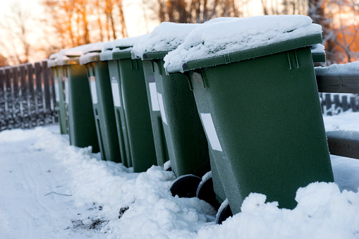 Green garbage cans in a row with snow on top on cold winter day with organge sunlight in background