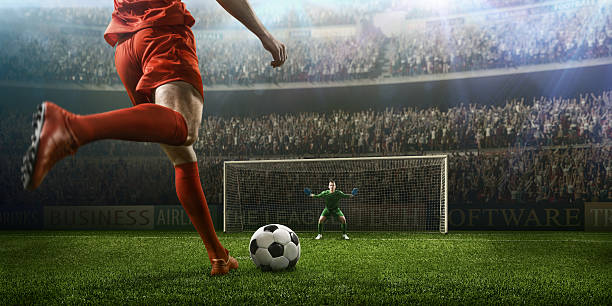 Soccer game moment with goalkeeper A male soccer player kicks a ball and soccer goalie jumping in motion for a ball while defending his gates on wide angle panoramic image of a outdoor soccer stadium or arena full of spectators under a sunny sky. The image has depth of field with the focus on the foreground part of the pitch. With intentional lensflares. Players are wearing unbranded soccer uniform. kicking stock pictures, royalty-free photos & images