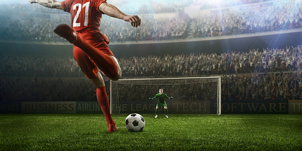 A male soccer player kicks a ball and soccer goalie jumping in motion for a ball while defending his gates on wide angle panoramic image of a outdoor soccer stadium or arena full of spectators under a sunny sky. The image has depth of field with the focus on the foreground part of the pitch. With intentional lensflares. Players are wearing unbranded soccer uniform.