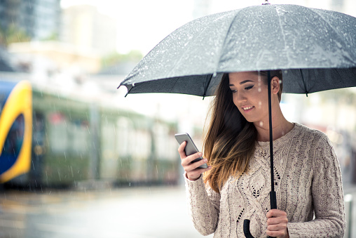 Woman standing on a street on a cold, rainy day. She is holding her mobile phone and ordering taxi online. In other hand she is holding an umbrella. She is smiling and looking at mobile phone. Tram in back, defocused. Shot in a city of Gold Coast, Australia.