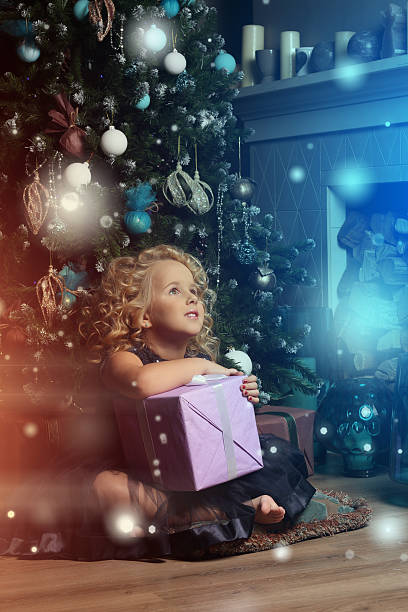 A cute little  blond girl like a doll with curly hair sitting with a gift  on the pillow near a fire-place and Christmas tree. New year waiting and celebration concept