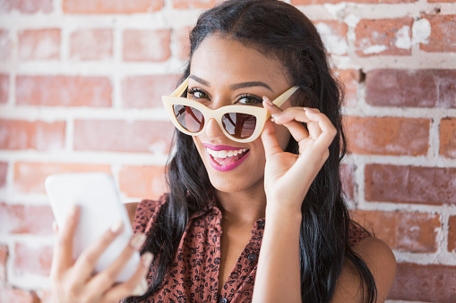 Head and shoulders of a beautiful young woman, mixed race African ethnicity and Caucasian, holding a mobile phone and looking over her sunglasses at the camera. She is standing in front of a brick wall.