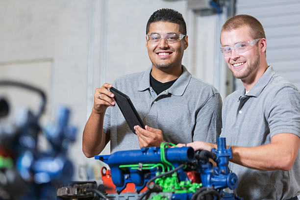 Two multi-racial men in auto mechanic school with engine Two multi-ethnic young men in vocational school, taking a class on reparing diesel engines.  They are working on an engine that has had parts painted different colors for training purposes.  They are wearing safety glasses. The Hispanic man is holding a digital tablet. multiengine stock pictures, royalty-free photos & images