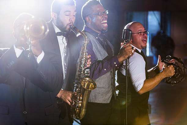 Jazz band performing at a nightclub A jazz band performing at a nightclub.  The four male musicians are standing together, playing a trumpet, saxophone, and tambourine.  A mature, African American man is the lead singer, singing into a microphone. jazz music stock pictures, royalty-free photos & images