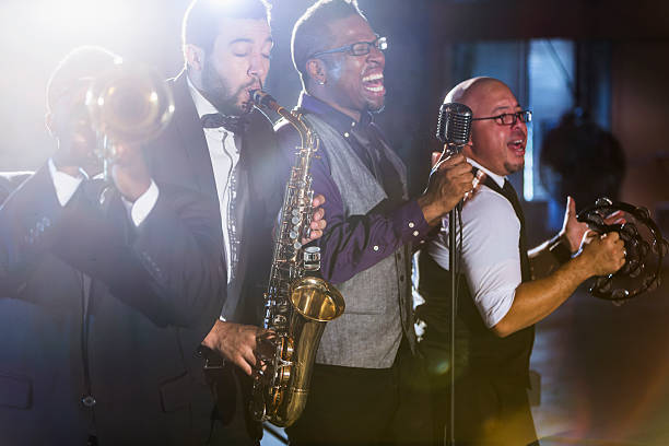 Jazz band performing at a nightclub A jazz band performing at a nightclub. The four male musicians are standing together, playing a trumpet, saxophone, and tambourine. A mature, African American man is the lead singer, singing into a microphone. performance group stock pictures, royalty-free photos & images
