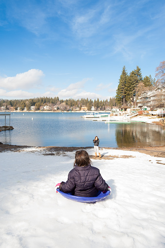 A pretty girl has fun in the snow as she rides her sled down a hill. She's dressed warmly. Shot from behind her, a beautiful lake and trees can be seen as her mother is taking a photo of her with her mobile device.  Plenty of room for copy. Shot at Lake Arrowhead, CA USA.