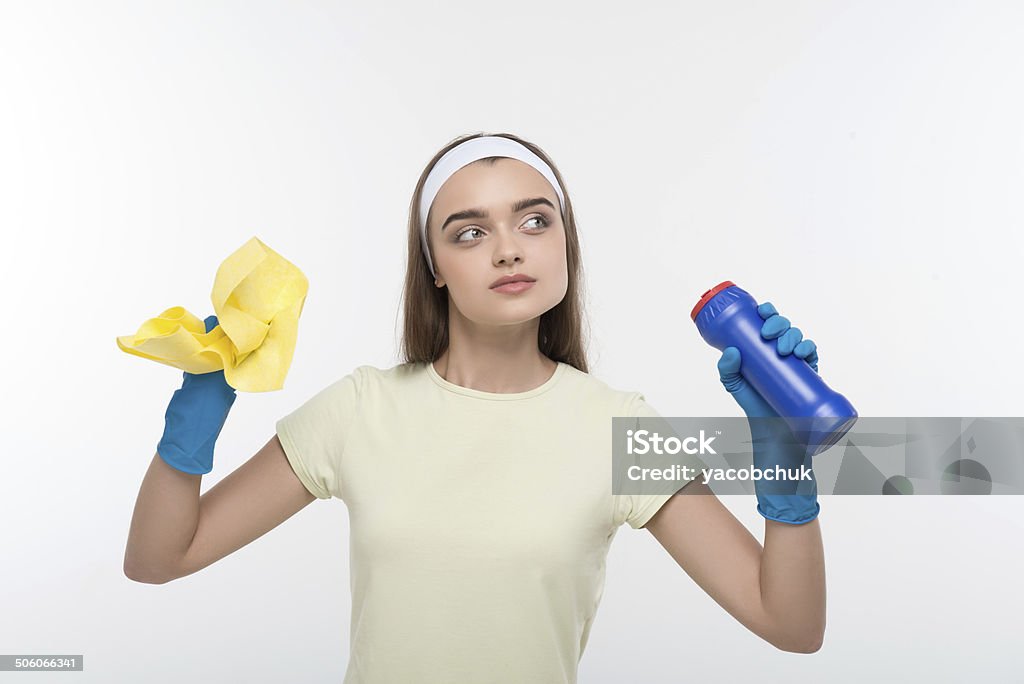 Maid and household chores Half-length portrait of pretty young dark-haired housemaid wearing white shirt holding abstergent and duster thinking about the rest. Isolated on white background Adult Stock Photo