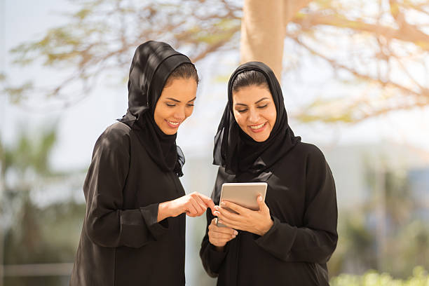 zwei emirati-frauen, die diskussion mit einem digitalen tablet - zurückhaltende kleidung stock-fotos und bilder