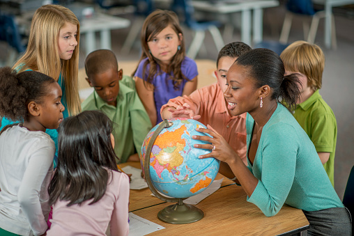 A multi-ethnic group of elementary age students are learning the places of the world by looking at a globe in geography and history class.