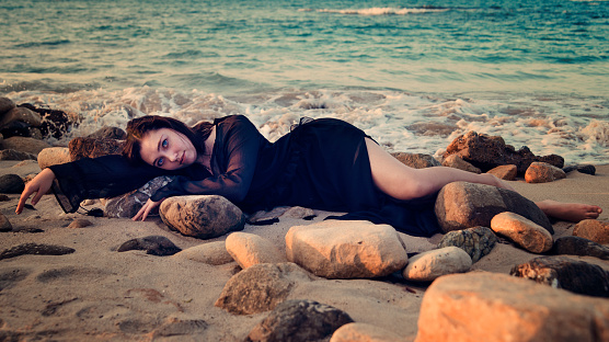 Mystical young woman lying down on rocks at the beach, at sunset hour, connecting with nature. She is wearing a long transparent black dress over a black tube dress. This was shot at Puerto Plata in Dominican Republic. Horizontal full length outdoors shot with copy space.
