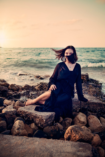 Mystical young woman sitting on rocks at the beach, at sunset hour, connecting with nature. She is wearing a long transparent black dress over a black tube dress. This was shot at Puerto Plata in Dominican Republic. Vertical full length outdoors shot with copy space.