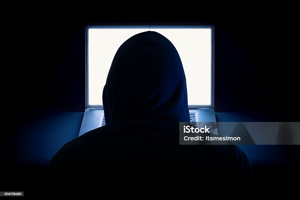 Hacker using laptop A hacker wearing a hoodie using a laptop in the dark. Computer Stock Photo