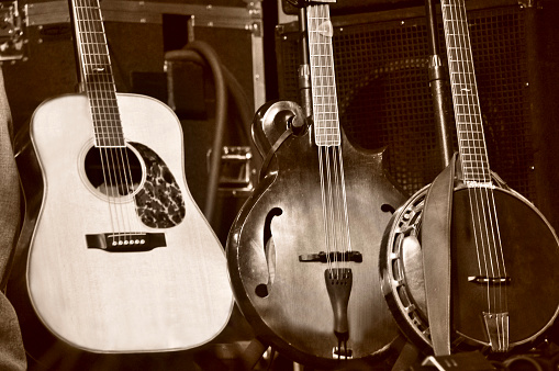 A medium closeup of three stringed folk instruments on a stage - a guitar (6-string), a bass mandolin (bouzouki), and a Banjo (5-string). The image is a brown monochrome. An amp and other audio equipment sits behind the instruments. The Banjo has a leather strap draped over it.