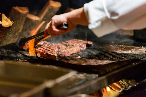 A group of flat iron steaks cooking on a grill over a live fire.  A chef is using tongs to turn the steaks.