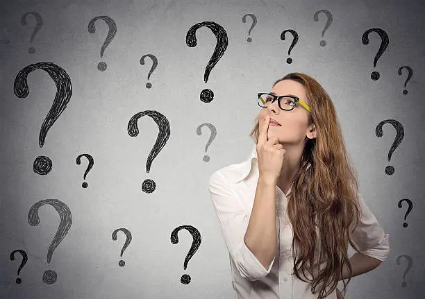 Thinking business woman with glasses looking up at many question marks isolated on gray wall background