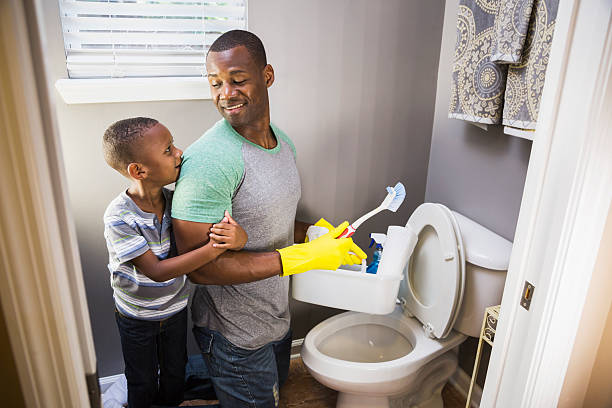 African American man with son, cleaning bathroom toilet An African American man doing housework.  He is kneeling in the bathroom at the toilet bowl, wearing rubber gloves, holding cleaning supplies.  His little boy is keeping him company, hugging his arm. role reversal stock pictures, royalty-free photos & images