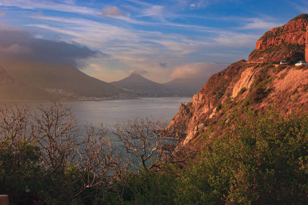 Sunset at Hout Bay in the Western Cape Province of South Africa.  The rays of the setting sun hit the mountain side and bring out the dramatic colours of the cliffs.  A couple of cars are seen driving uphill. Photo shot in the Blue Hour in the horizontal format; copy space.