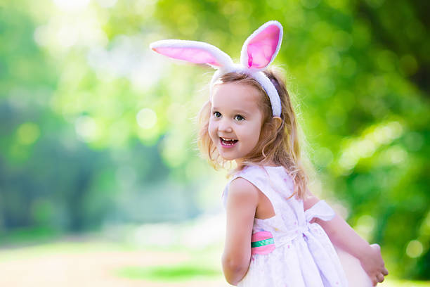 Little girl having fun on Easter egg hunt. Kids in bunny ears and rabbit costume. Children searching for eggs in the garden. Toddler kid playing outdoor. Child laughing and smiling on a spring day