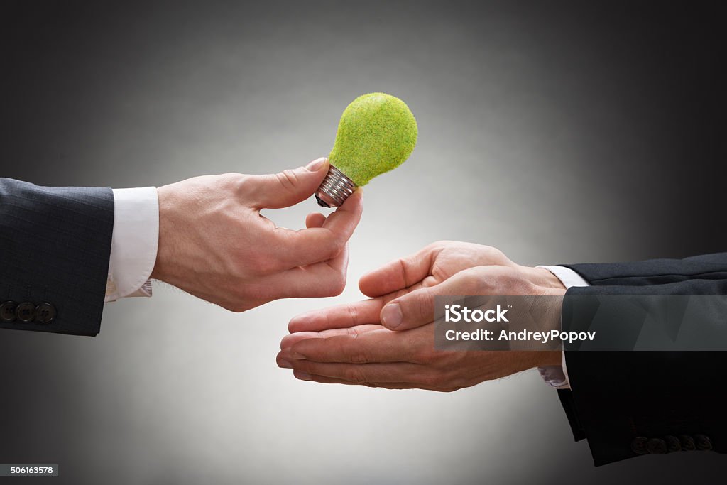 Businessman Giving Eco Light Bulb To Other Businessperson Close-up Of Businessman Hand Giving Eco Light Bulb To Other Businessperson Light Bulb Stock Photo