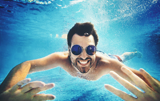 Closeup of smiling man diving in a swimming pool. He's moving towards camera very close to the bottom. Wearing sunglasses, looking at camera and waving.