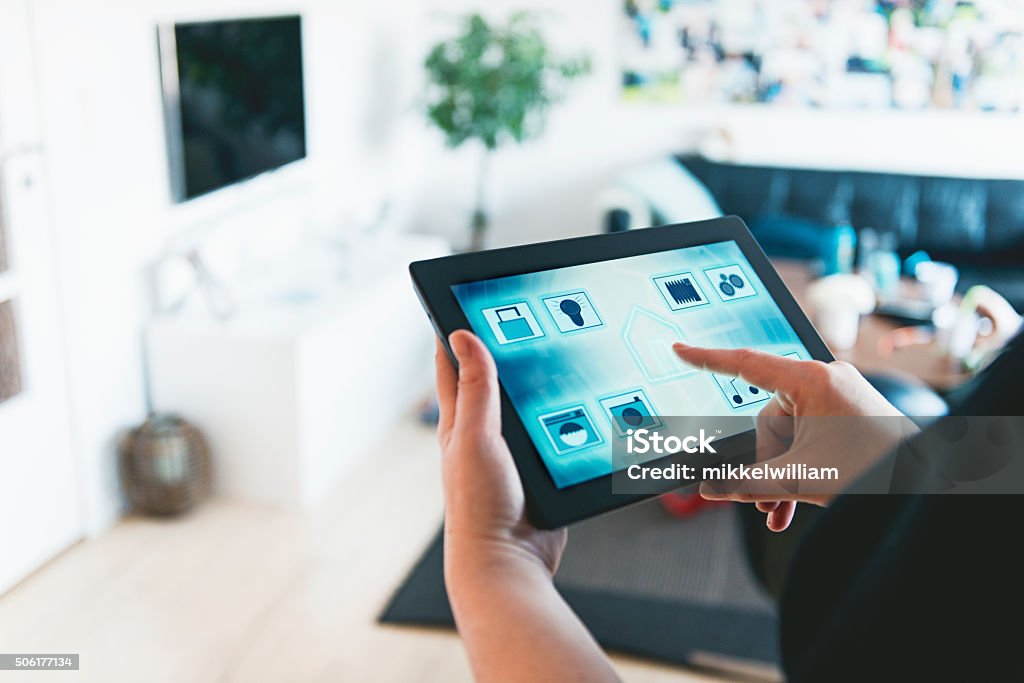 Smart home automation and control with digital tablet Tablet with an app controls several features in the home such as door lock, lights, washing machine, surveillance camera, music, water use and more. Woman stands in a living room and press the center of the tablet with an icon of a house.  Home Automation Stock Photo