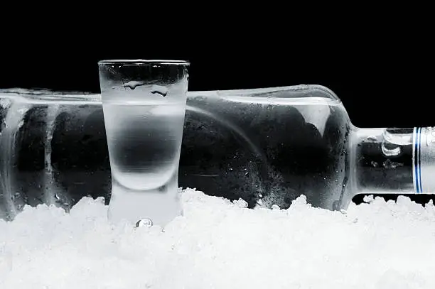 Studio shot of bottle with glass of vodka lying on ice on black background