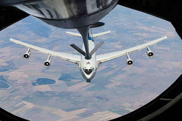 A small white jet plane flies high in a clear blue sky, surrounded by soft, fluffy clouds. The aircraft is seen from below.