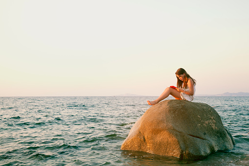 Twelve years old girl reading a book sitting on a boulder in the sea.