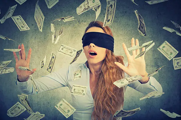 Blindfolded young entrepreneur businesswoman trying to catch dollar bills banknotes flying in the air on gray wall background. Financial corporate success or crisis challenge concept