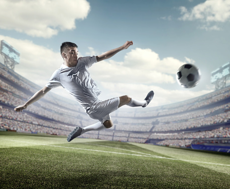 A male soccer player makes a dramatic play by jumping horizontally. He attempts to kick the ball with his feet. The stadium is blurred behind him. It's sunny weather.